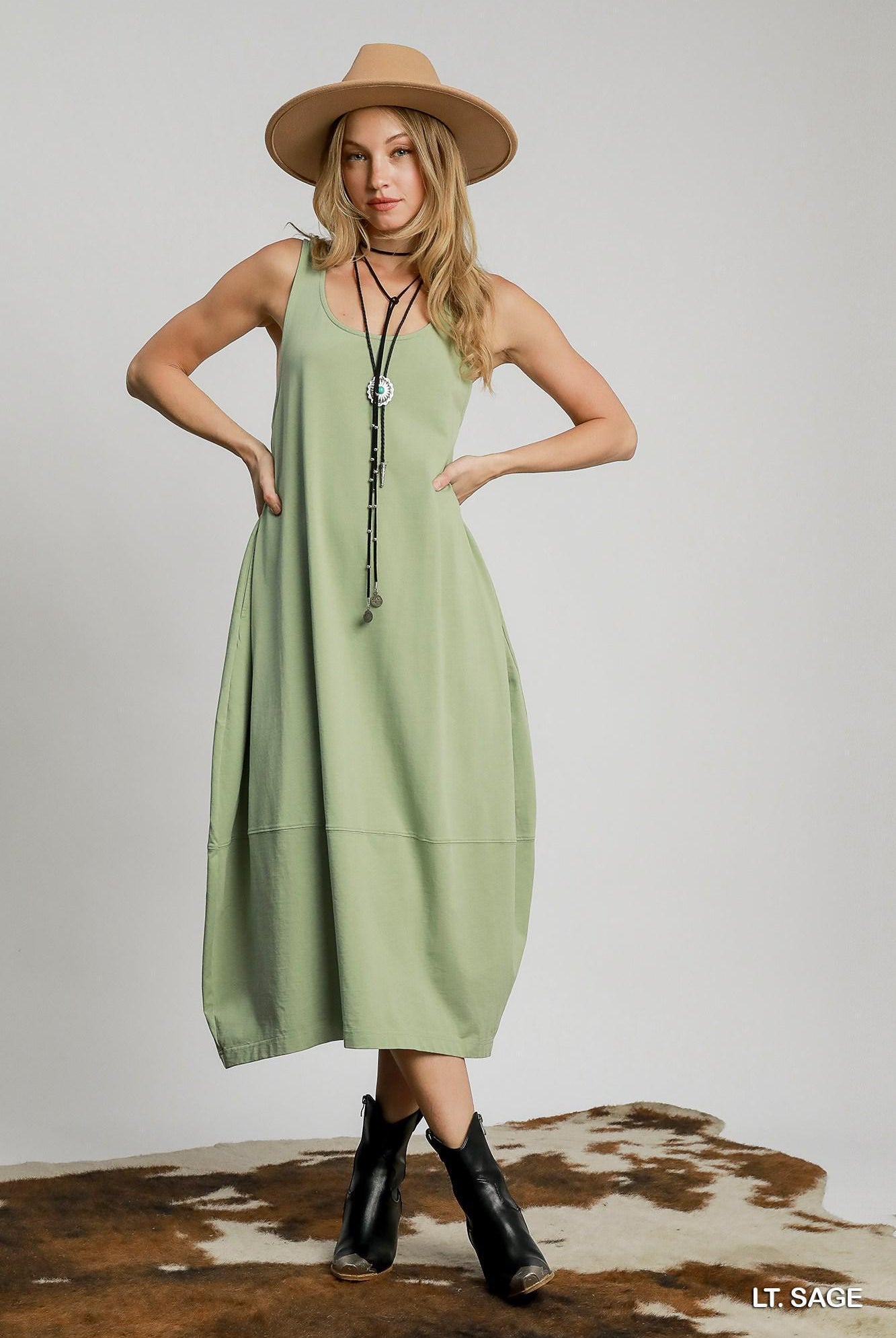 Woman wearing a light sage dress with a hat and boots on a plain background