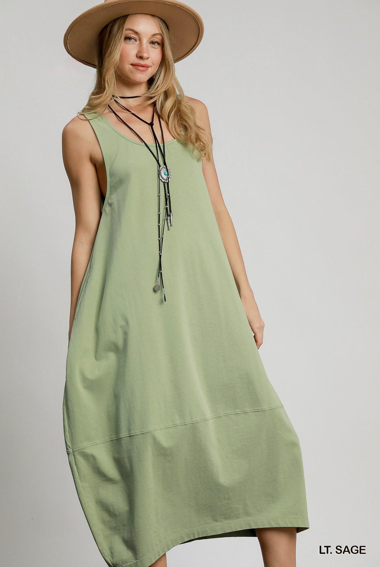 A woman wearing a sleeveless sage green midi dress with a relaxed, loose fit, styled with a wide-brimmed tan hat and layered necklaces, set against a plain light gray studio background.
