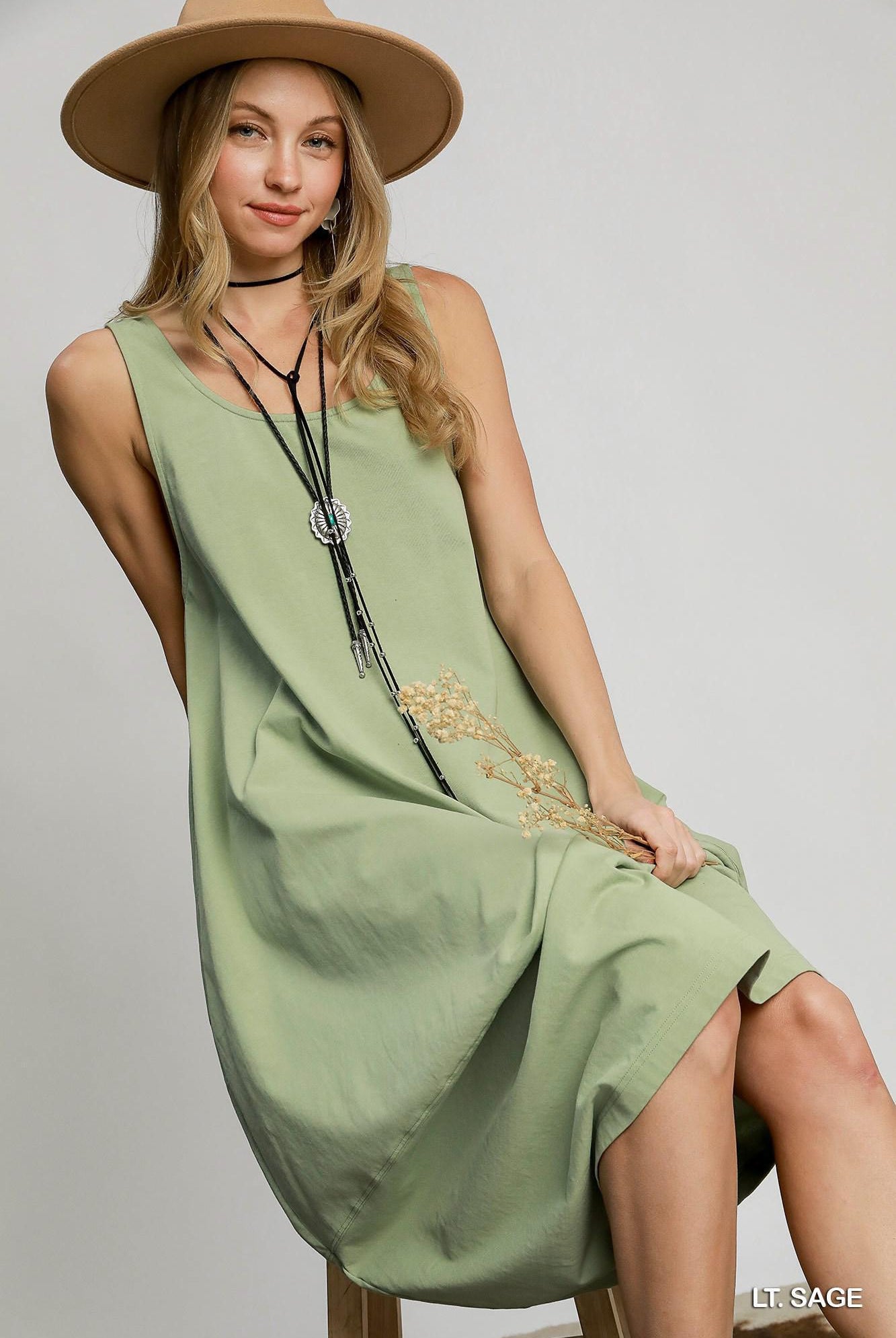 A woman seated on a stool wearing a sleeveless sage green dress with gold embroidered detailing near the hem, a wide-brim tan hat, and long black necklaces, photographed against a light gray studio background.