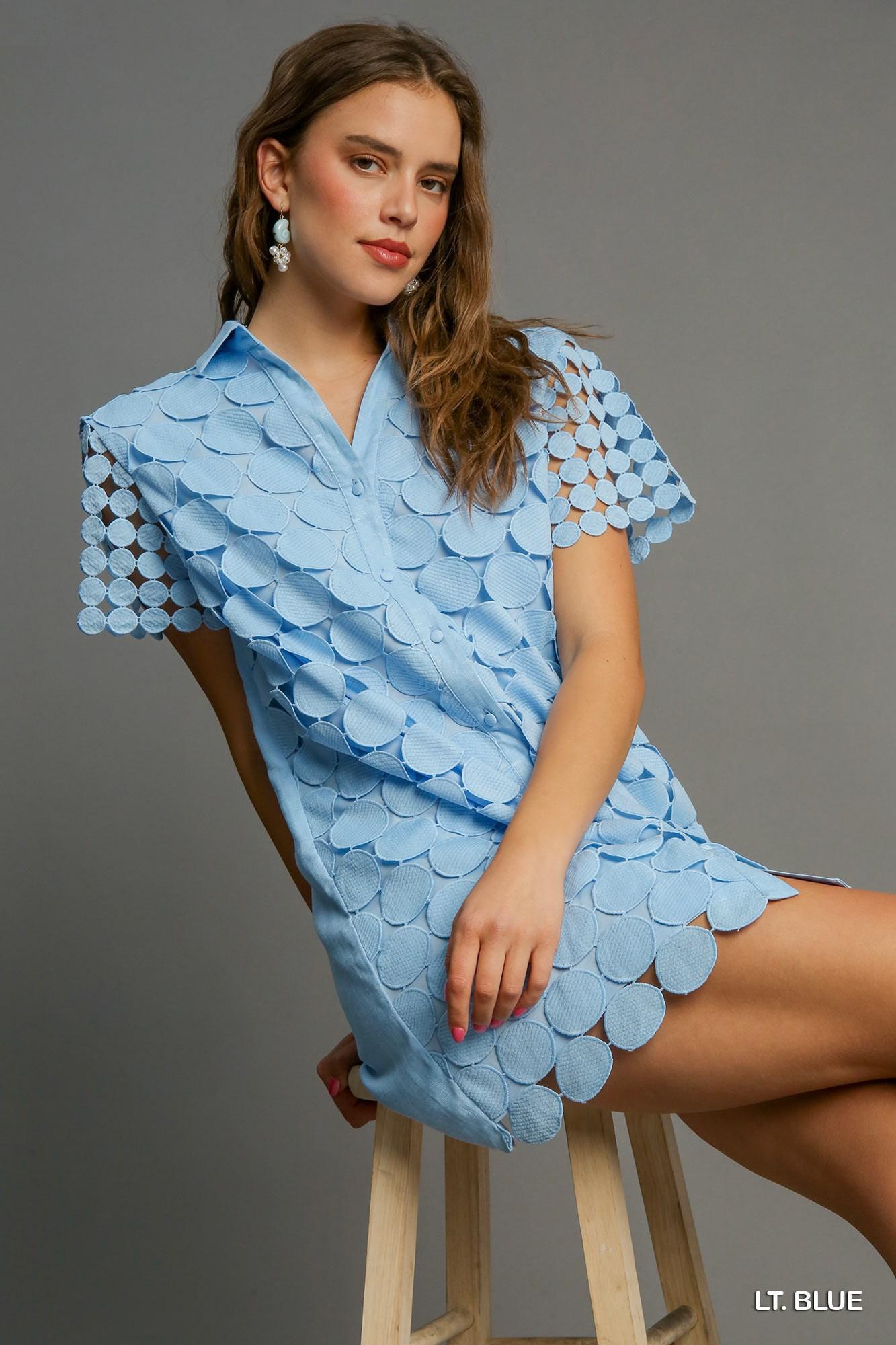 Model wearing light blue shirt dress with circular appliqué detailing and short sleeves, seated