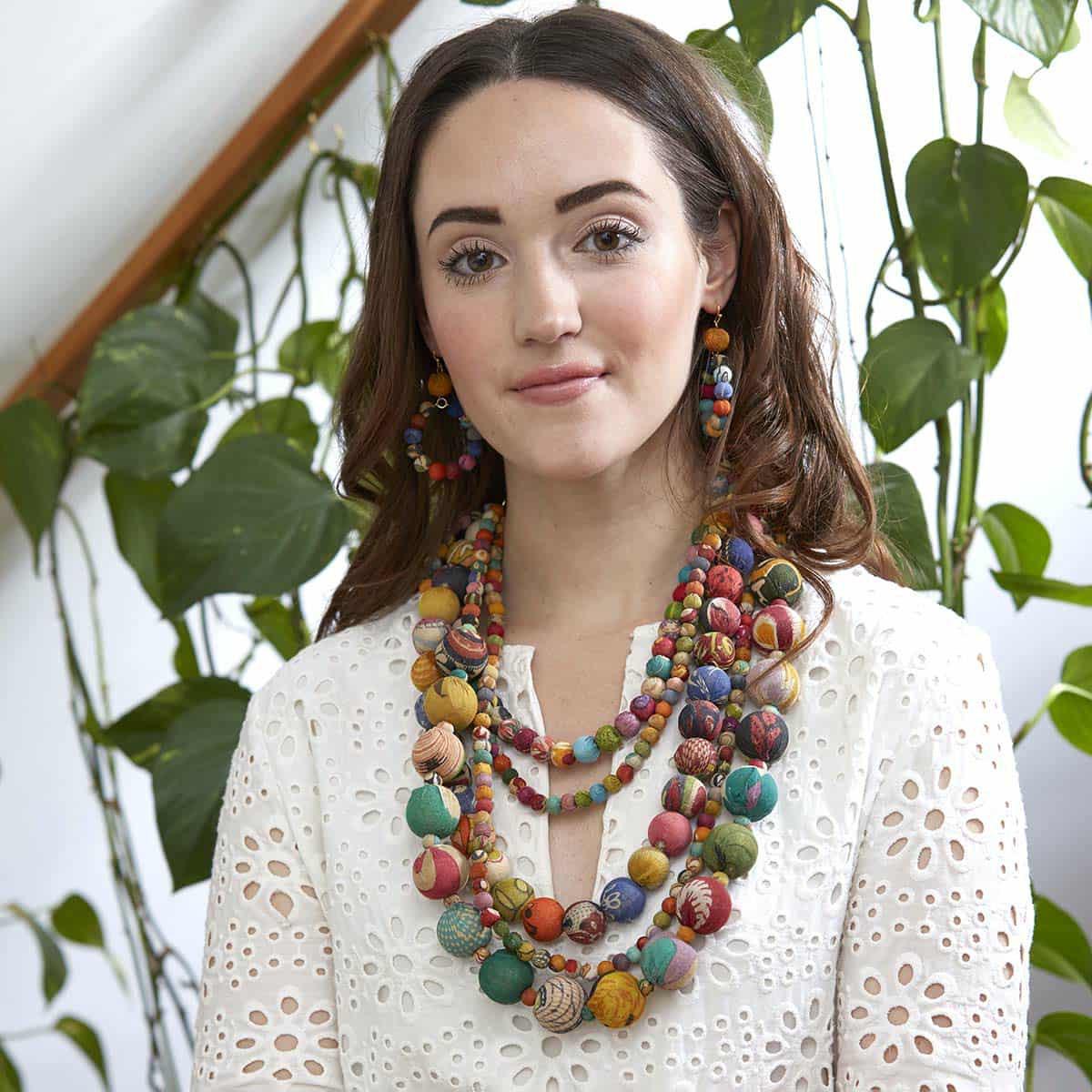 Woman wearing a colorful beaded necklace and earrings with plants in the background.WorldFinds multicolor fabric bead statement necklace handmade from recycled Kantha textiles – vibrant fair-trade jewelry