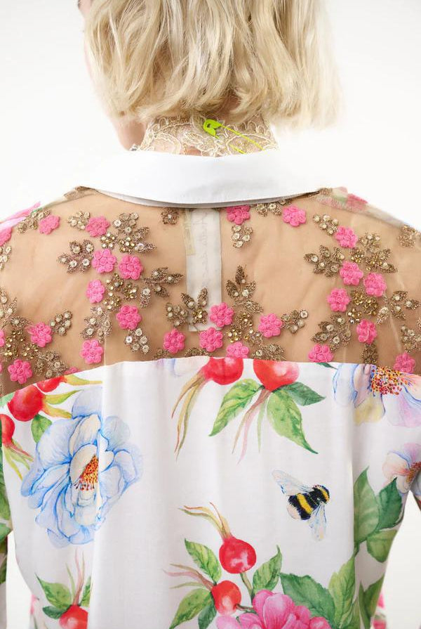 Back view of a white blouse with a colorful floral print and pink floral embroidery on a sheer mesh yoke, worn by a model.