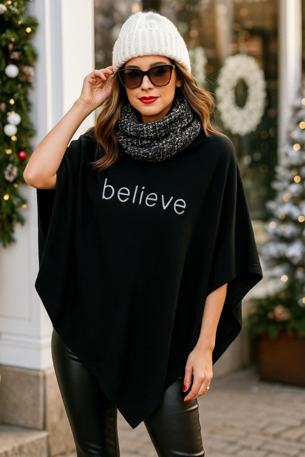 Woman wearing a black lightweight poncho with the word “believe” in rhinestones, styled for the winter holidays with a knit beanie and leather leggings.
