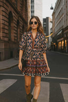 Urban boho chic woman wearing a THML geometric print mini dress with puff sleeves and tiered hem, styled with ankle boots on a city street.