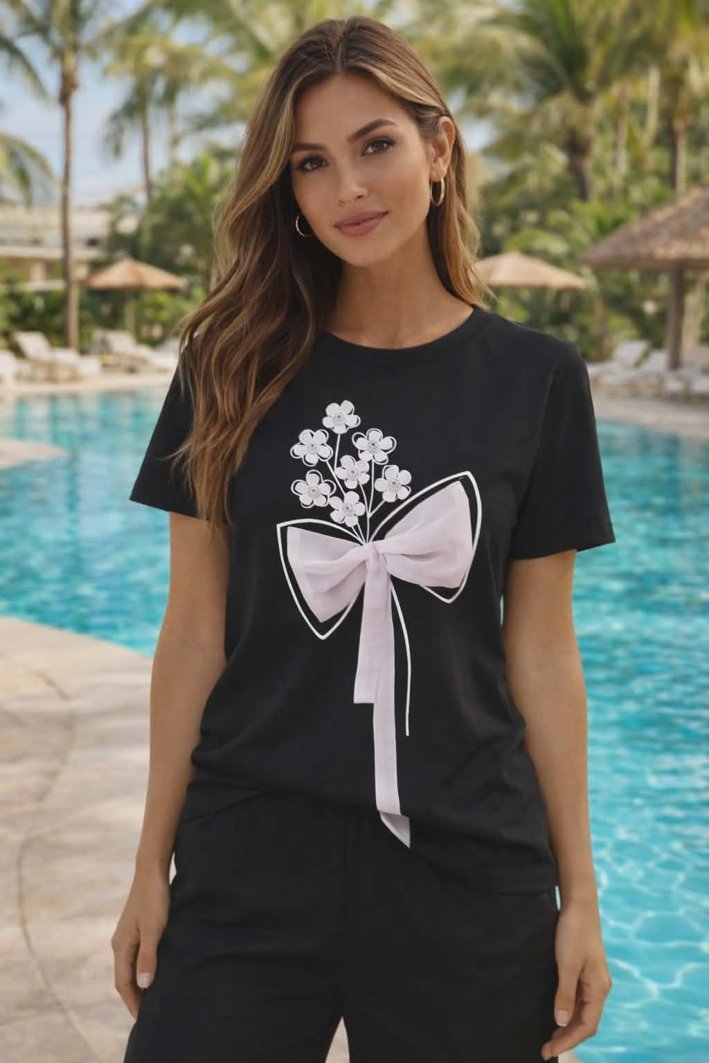 Black Michael Tyler women’s tee featuring a soft pink bow graphic with floral appliqués and rhinestone details styled with black pants in a resort setting
