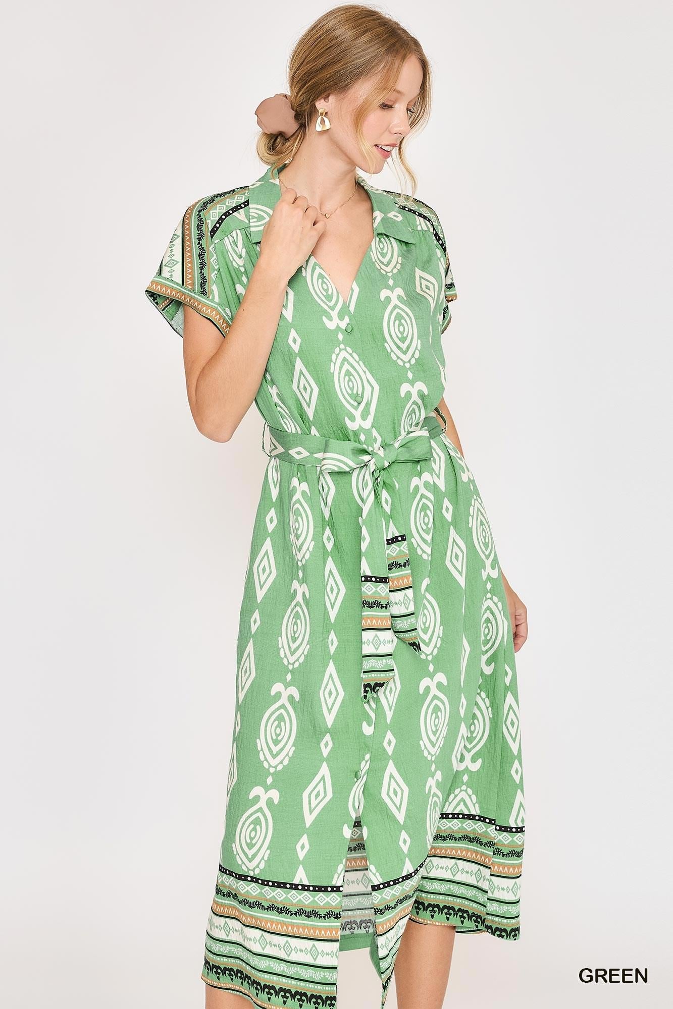 A woman modeling a green wrap-style midi dress with a white geometric pattern and a tie waist, photographed against a plain light background.