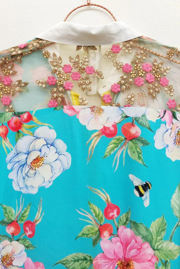 A close-up of a floral printed garment, likely a blouse or dress, featuring a bright turquoise background with large pink, white, and blue flowers, green leaves, red berries, and a small bumblebee motif. The upper portion includes sheer mesh with pink floral embroidery and gold beadwork over a white collar.
