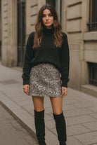 Woman wearing a black sweater, sequin skirt, and knee-high boots on a city street.