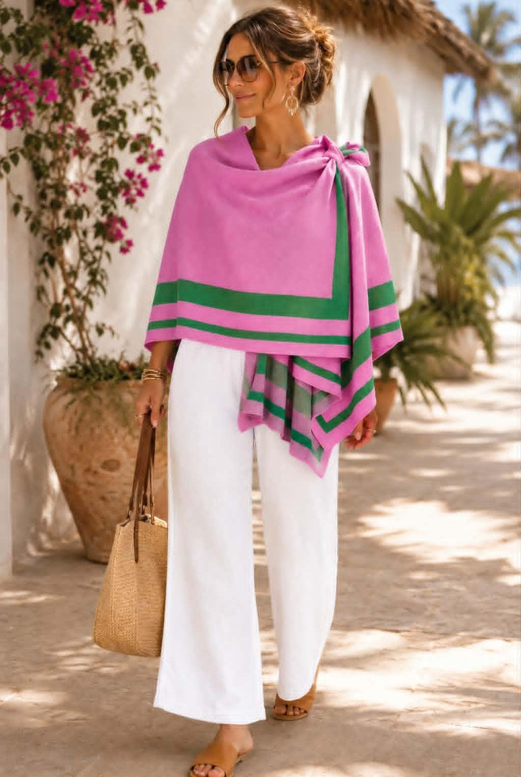 A woman wearing a bright pink wrap or poncho with green striped borders, styled with white wide-leg pants, sunglasses, and a straw tote bag in an outdoor sunlit setting.