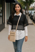 Woman walking on a sidewalk wearing a black top, white shirt, blue jeans, and carrying a beige crossbody bag.