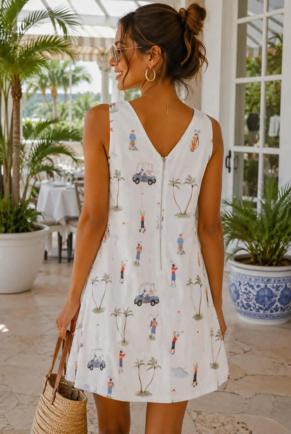 A woman wearing a sleeveless white V-neck dress with a colorful golf-themed print, standing on a patio and holding a straw bag.