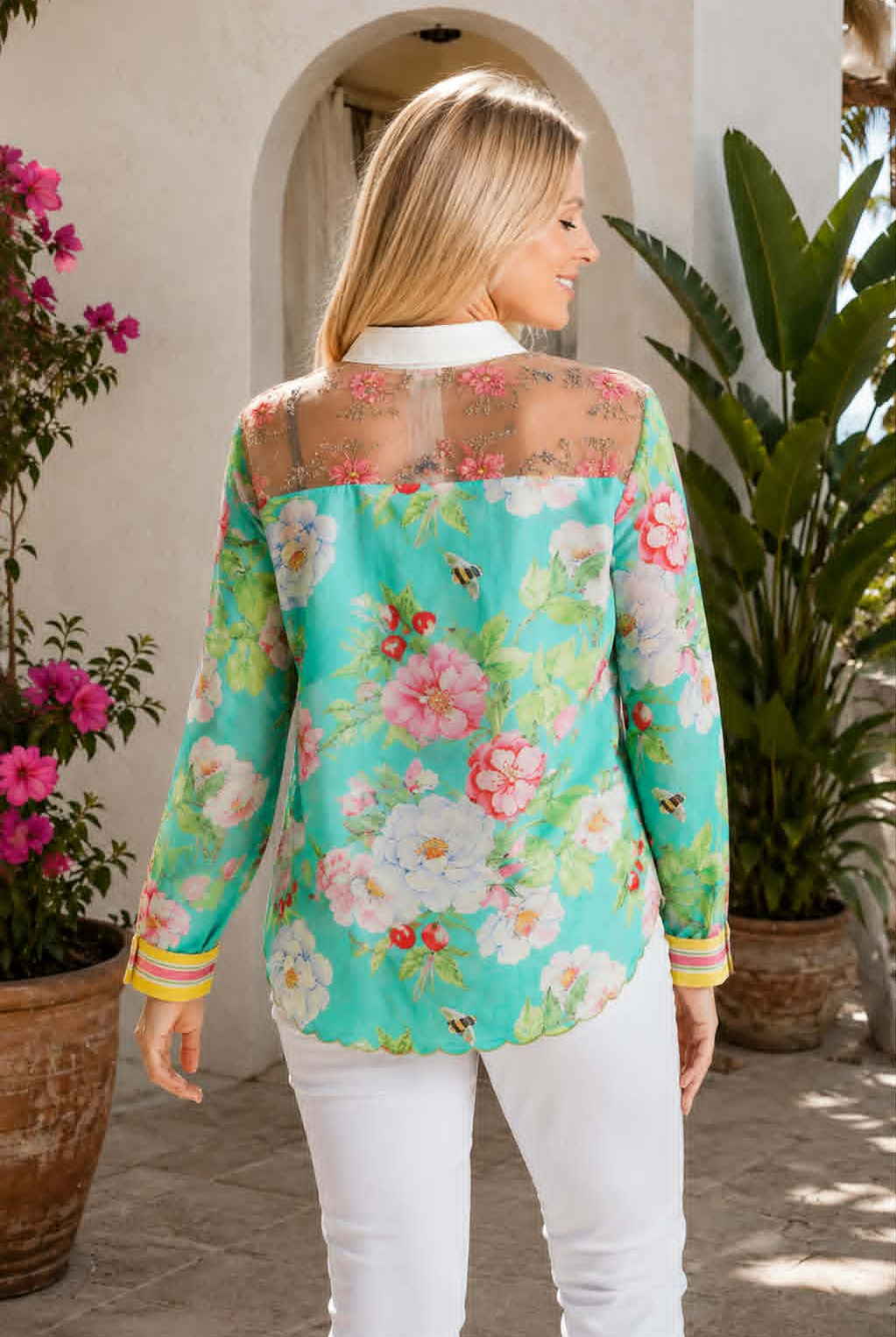 A woman wearing a turquoise floral long-sleeve blouse with sheer mesh shoulders and white pants, photographed outdoors in front of a stucco archway with potted plants.