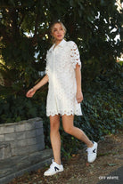 Model wearing off-white floral lace shirt dress with short sleeves and scalloped hem, styled with white sneakers outdoors