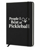 Black hardcover journal with "People I Beat at Pickleball" printed in white lettering, featuring pickleball paddle graphics, an elastic closure, and ribbon bookmark.