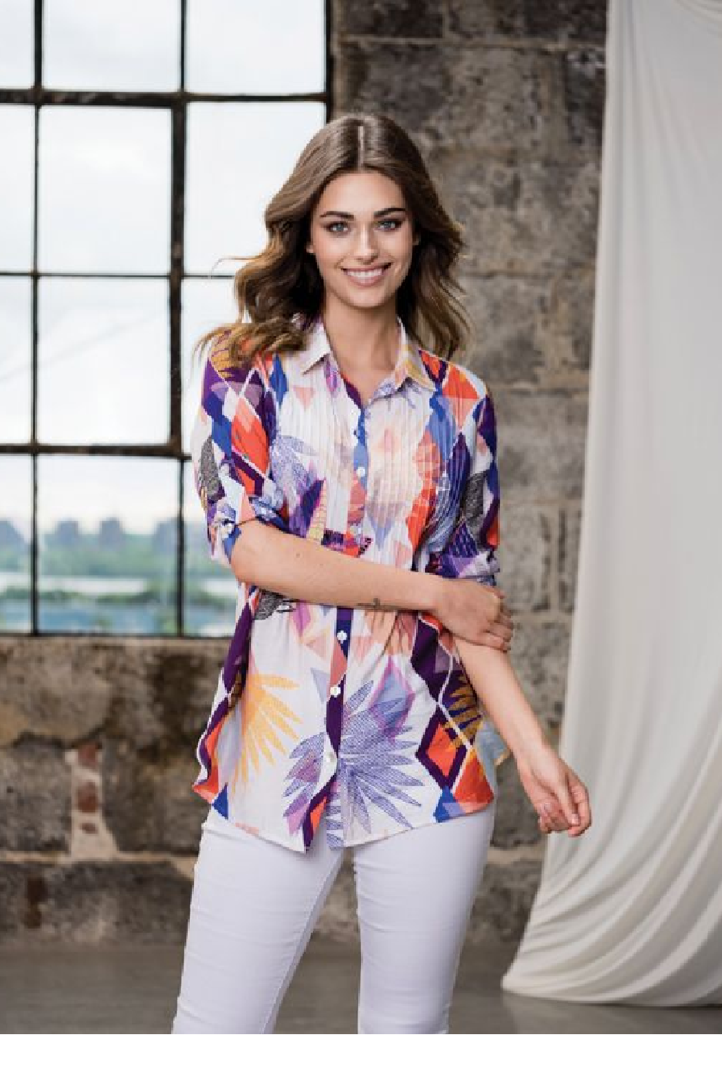 Model wearing Alison Sheri colorful geometric floral print button-down blouse with white pants.
