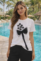 White Michael Tyler women’s tee featuring a black bow graphic with floral appliqués and rhinestone details styled with black pants in a resort setting