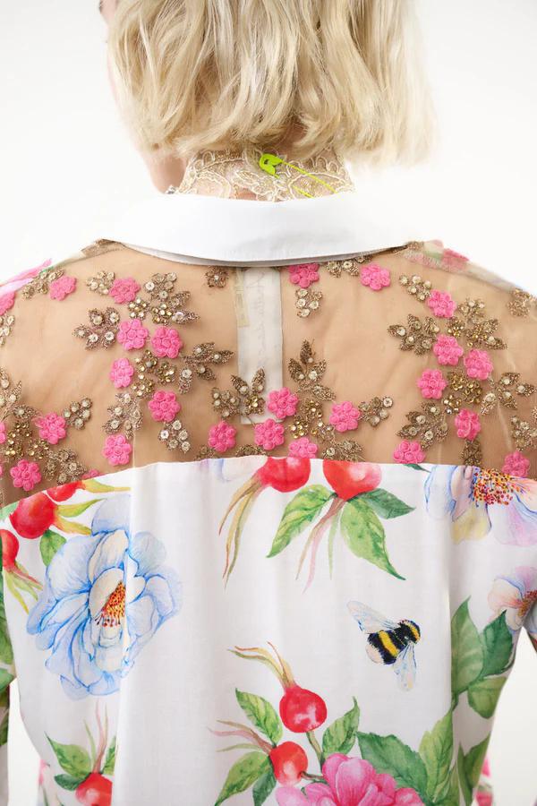 Back view of a white blouse with a colorful floral print and pink floral embroidery on a sheer mesh yoke, worn by a model.