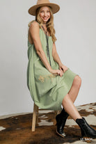 Woman wearing a light sage green dress with black boots and a beige hat, sitting on a wooden stool.