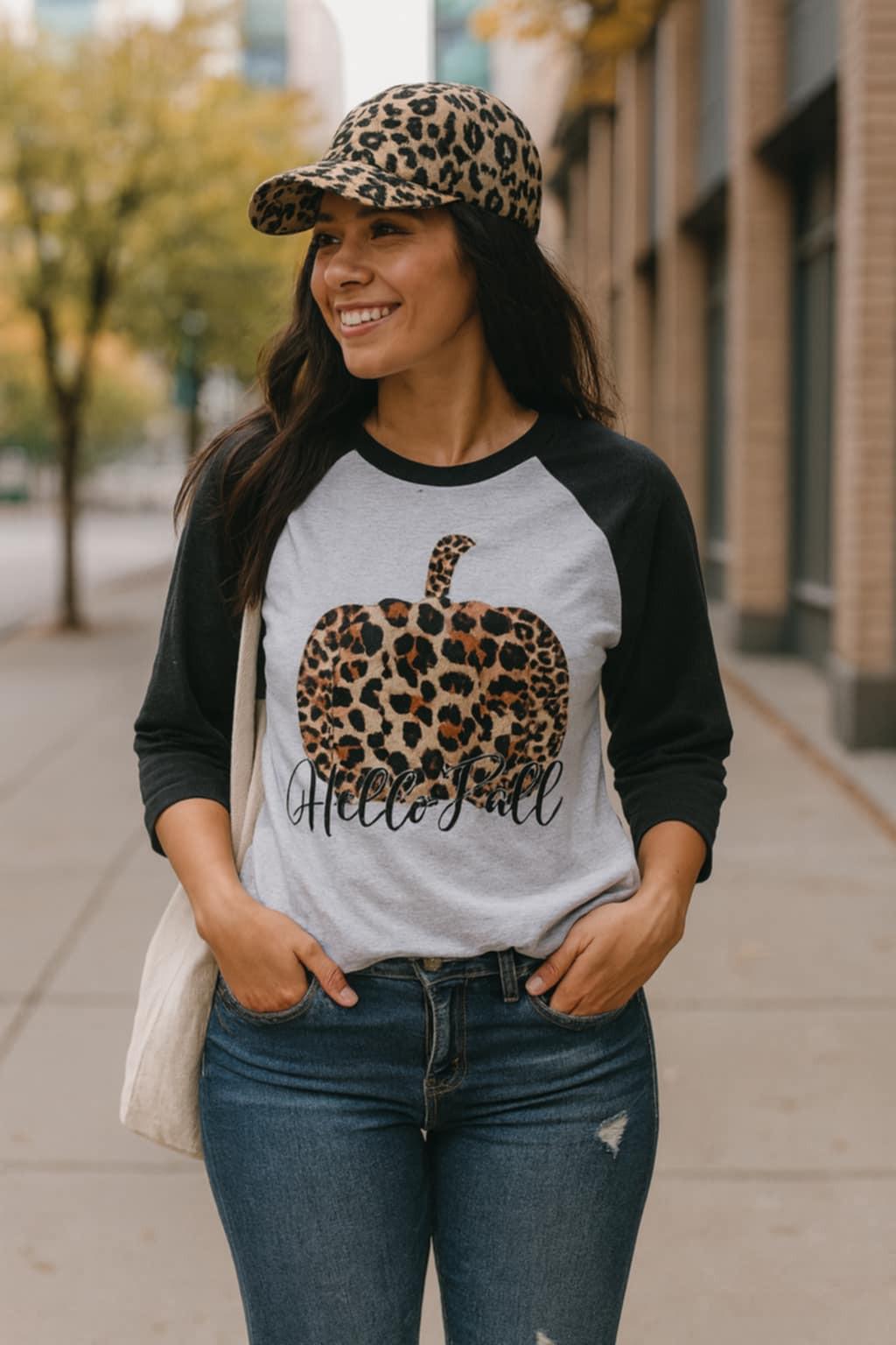 Urban chic woman wearing Hello Fall raglan tee with leopard pumpkin graphic, styled with jeans and leopard print cap.