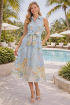 Model wearing a blue scenic print midi dress with tie waist and sleeveless button front by Entro, styled for a resort chic look