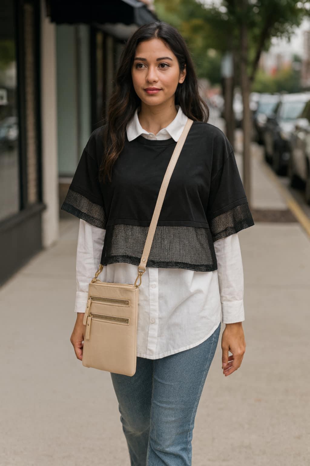Woman walking on a sidewalk wearing a black top, white shirt, blue jeans, and carrying a beige crossbody bag.