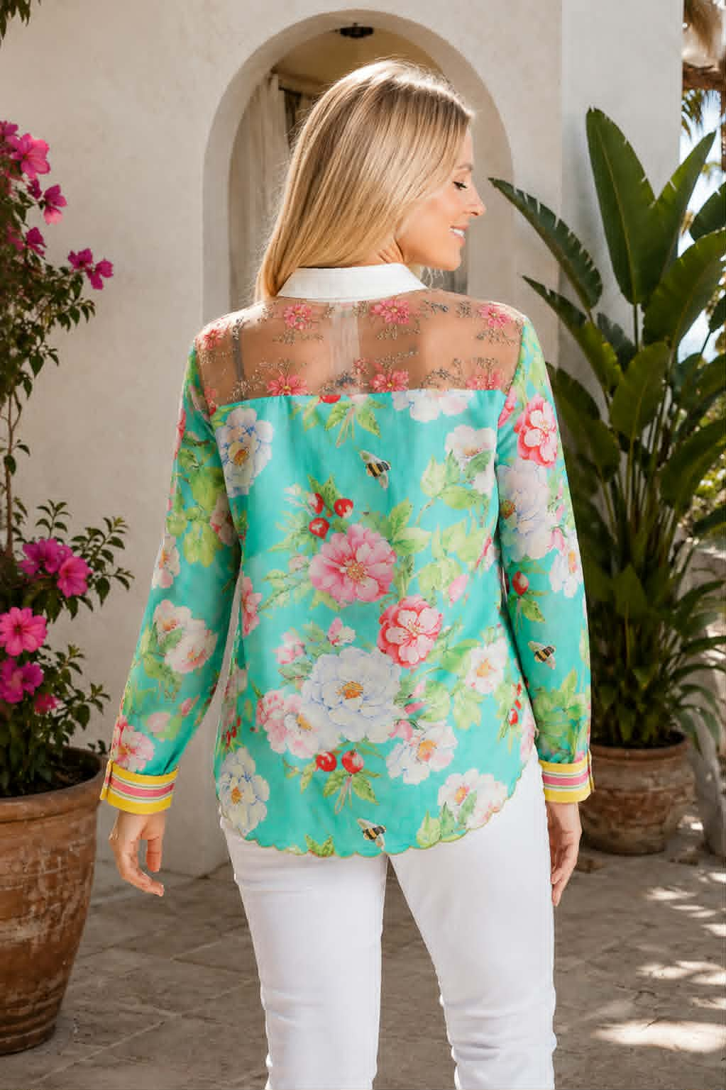 A woman wearing a turquoise floral long-sleeve blouse with sheer mesh shoulders and white pants, photographed outdoors in front of a stucco archway with potted plants.