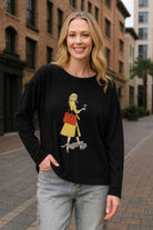 Model wearing Gabby Isabella black sweater featuring a colorful fashion-girl graphic with a yellow coat and red handbag, styled in an urban city setting — Petunias of Naples