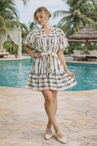 Umgee Off white mini dress with navy bow print, puff sleeves, and tie waist styled on a resort chic model by the pool